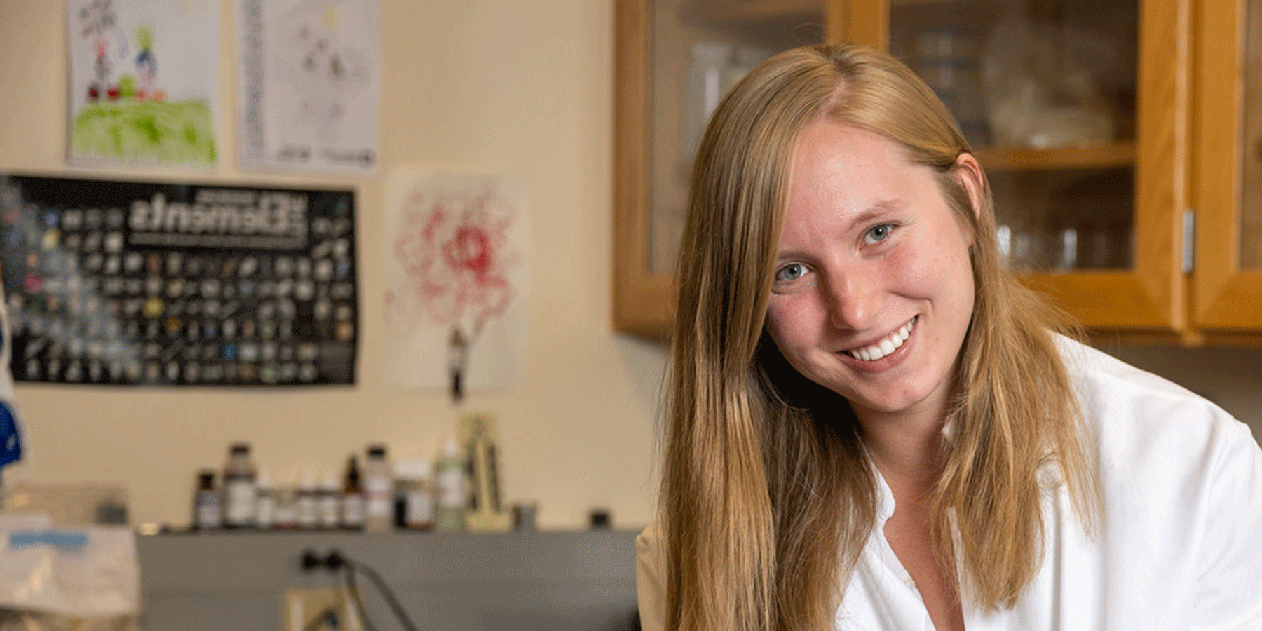 A white female student with long blond hair poses in a laboratory space. She wears a white lab coat. Behind her are brown cabinet doors, medical bottles, and various posters on the wall, including the Periodic Table of Elements.